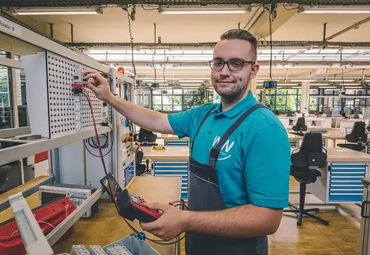 Bild mit einer Person in Arbeitskleidung stehend in einer Werkshalle Elektrotechnik