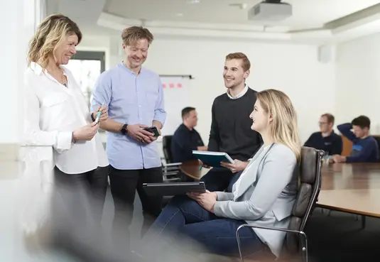 Bild mit vier Personen stehend und sitzend im Büro bei einer Besprechnung 