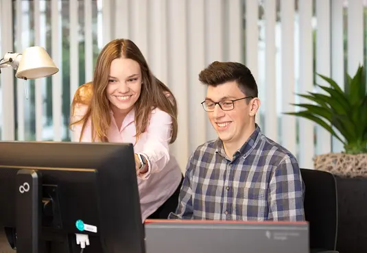 eine Frau stehend und ein Mann sitzend am Schreibtisch im Büro vor einem PC-Bildschirm