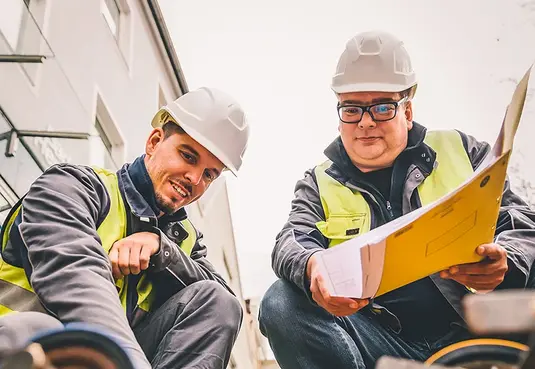 Bild mit zwei Personen kniend auf einer Baustelle in Arbeitskleidung mit Warnweste 